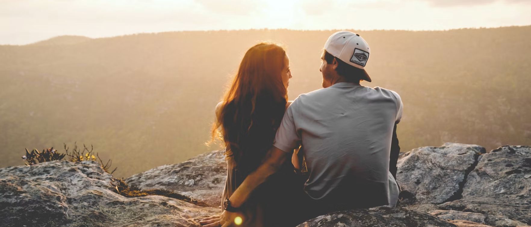 A couple sitting close together on a rocky outcrop, looking out over hills at sunset